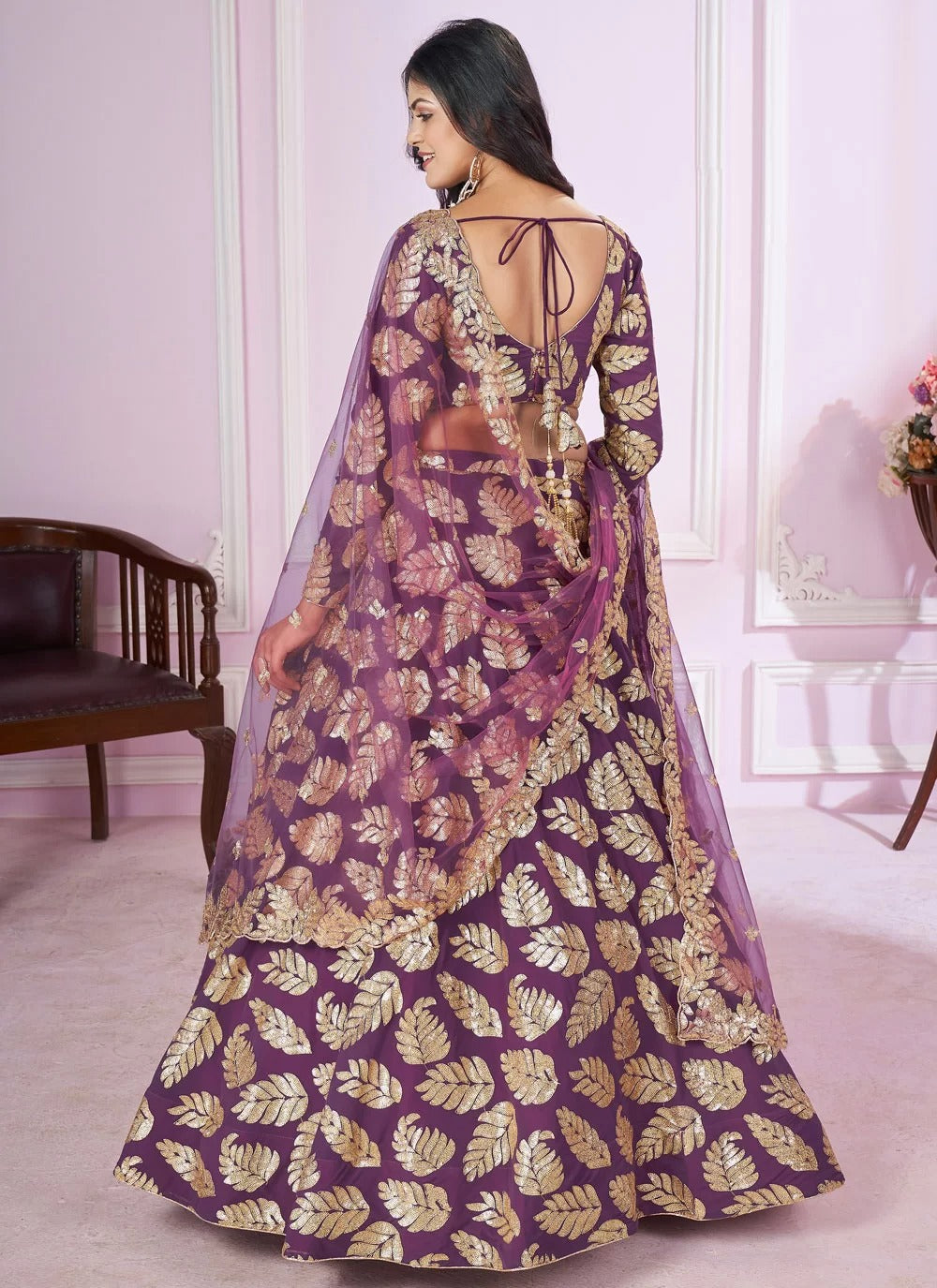 Woman wearing a purple and gold traditional outfit with a sheer dupatta in a room.