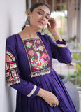 A woman wearing a purple embroidered Anarkali kurta with colorful floral and geometric patterns on the chest and sleeve, paired with silver earrings, in an outdoor setting.