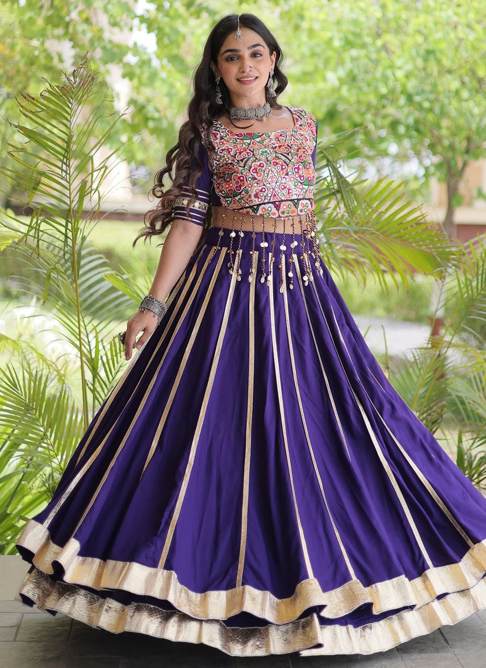 A woman wearing a purple traditional Indian lehenga choli with gold stripes and a floral embroidered bodice, standing outdoors in front of green foliage.
