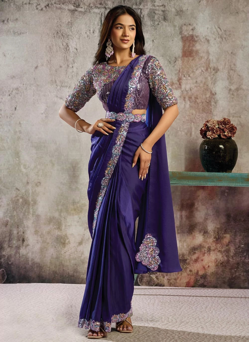 Woman wearing a purple saree with silver border against a textured wall.