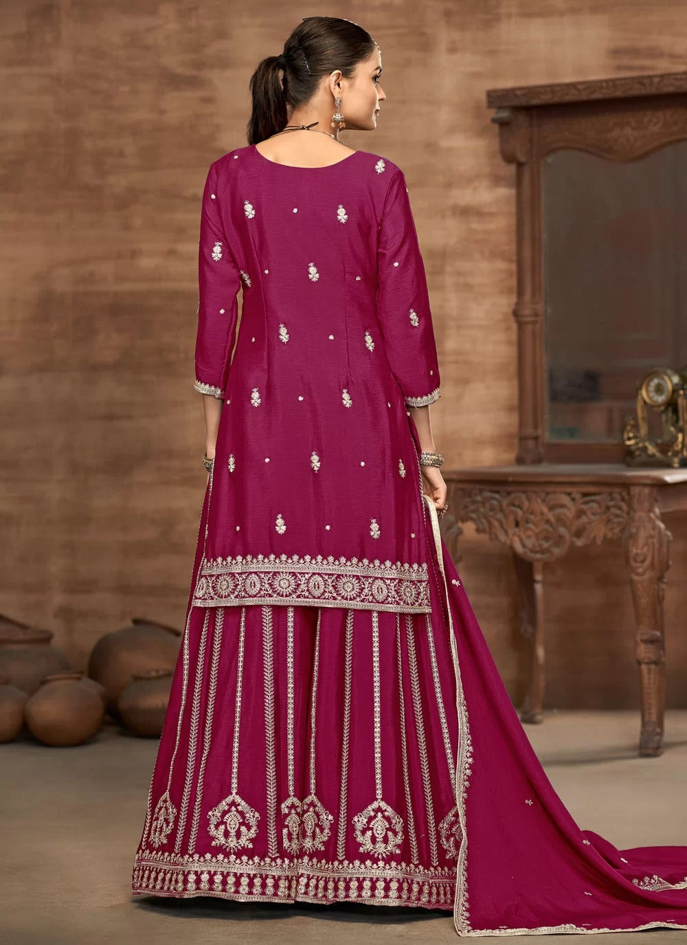 Woman wearing a magenta traditional outfit with white embroidery in a room with wooden furniture.