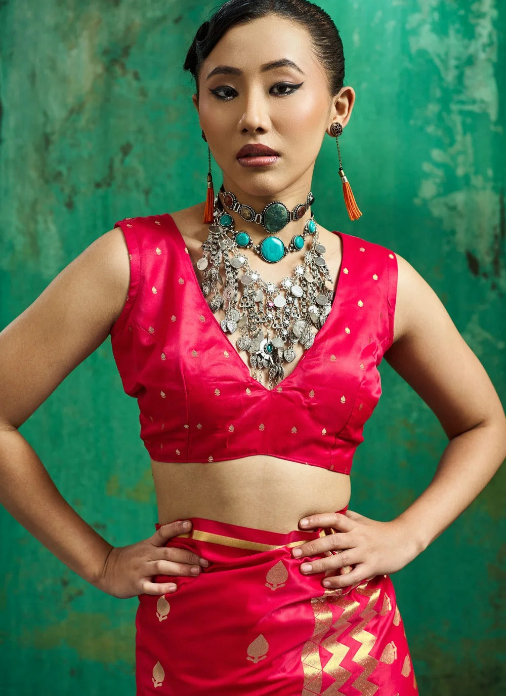 A woman modeling a bright pink traditional Indian outfit with gold zari detailing, featuring a sleeveless crop top and matching skirt, styled with a large silver necklace and turquoise earrings against a textured green backdrop.