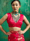 A woman modeling a bright pink traditional Indian outfit with gold zari detailing, featuring a sleeveless crop top and matching skirt, styled with a large silver necklace and turquoise earrings against a textured green backdrop.