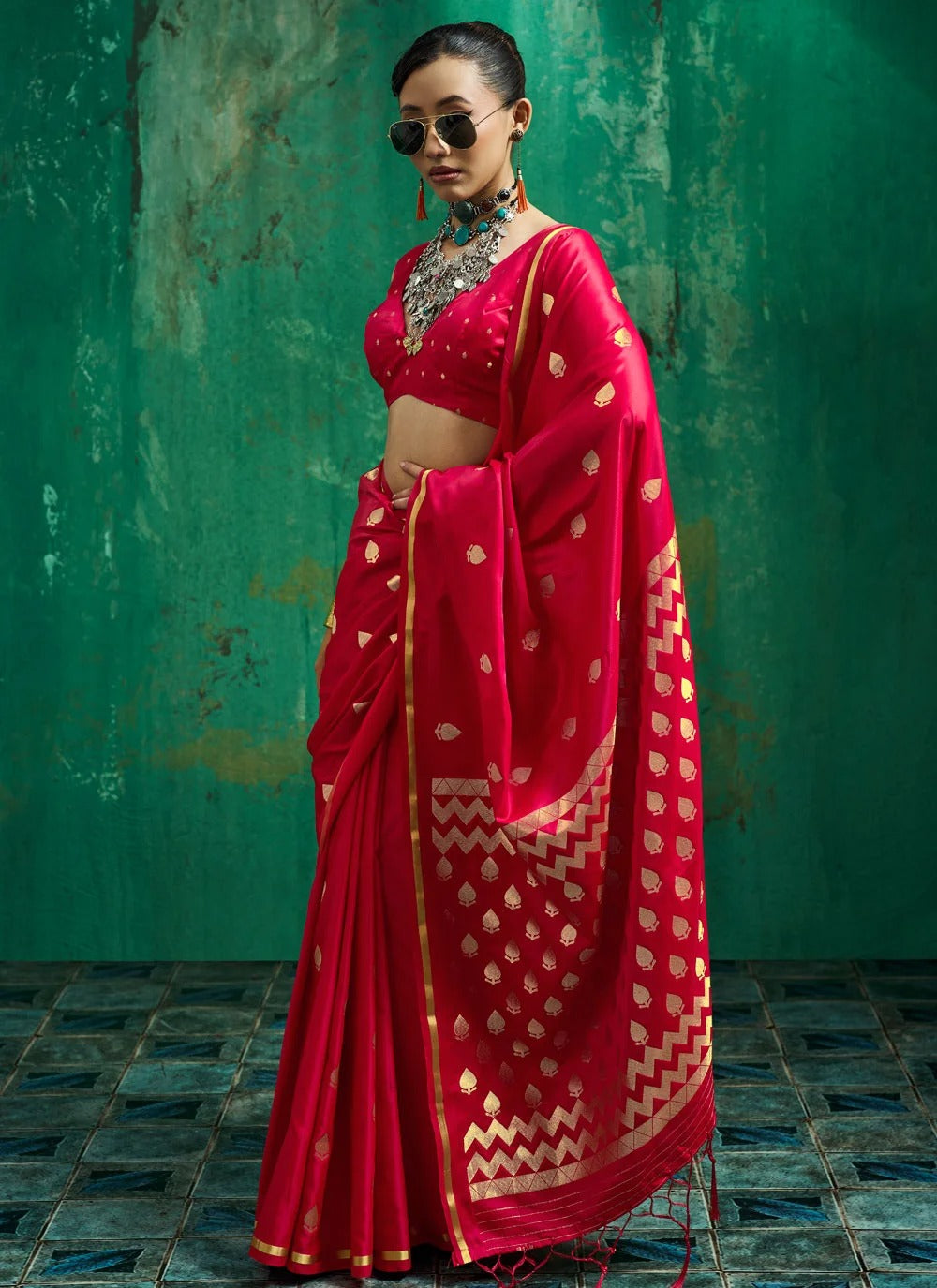 A woman models a bright red saree with gold zari woven motifs and borders, paired with a matching red blouse, set against a textured green wall.