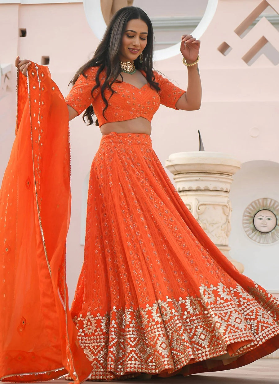 Woman in an orange traditional outfit with a matching dupatta, standing indoors.