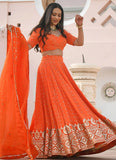 Woman in an orange traditional outfit with a matching dupatta, standing indoors.