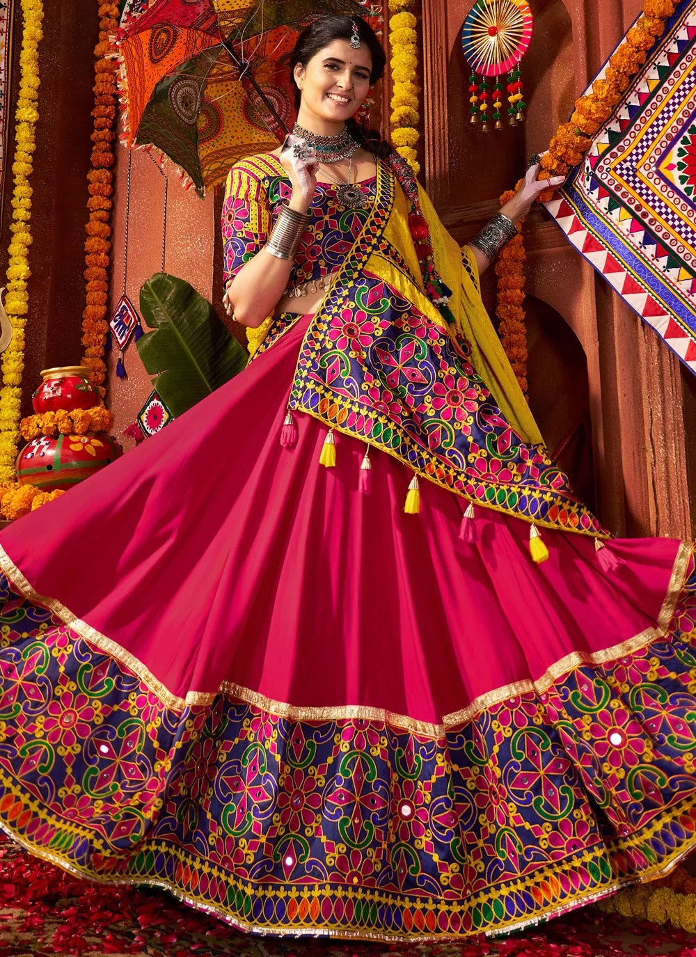 Woman in a colorful traditional outfit with intricate patterns, standing against a decorated background.