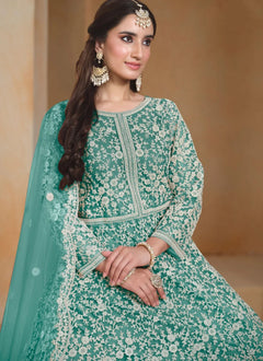 A woman wearing a mint green traditional embroidered outfit with white floral lace patterns, a matching dupatta, and ornate gold jewelry, posed against a warm beige background.