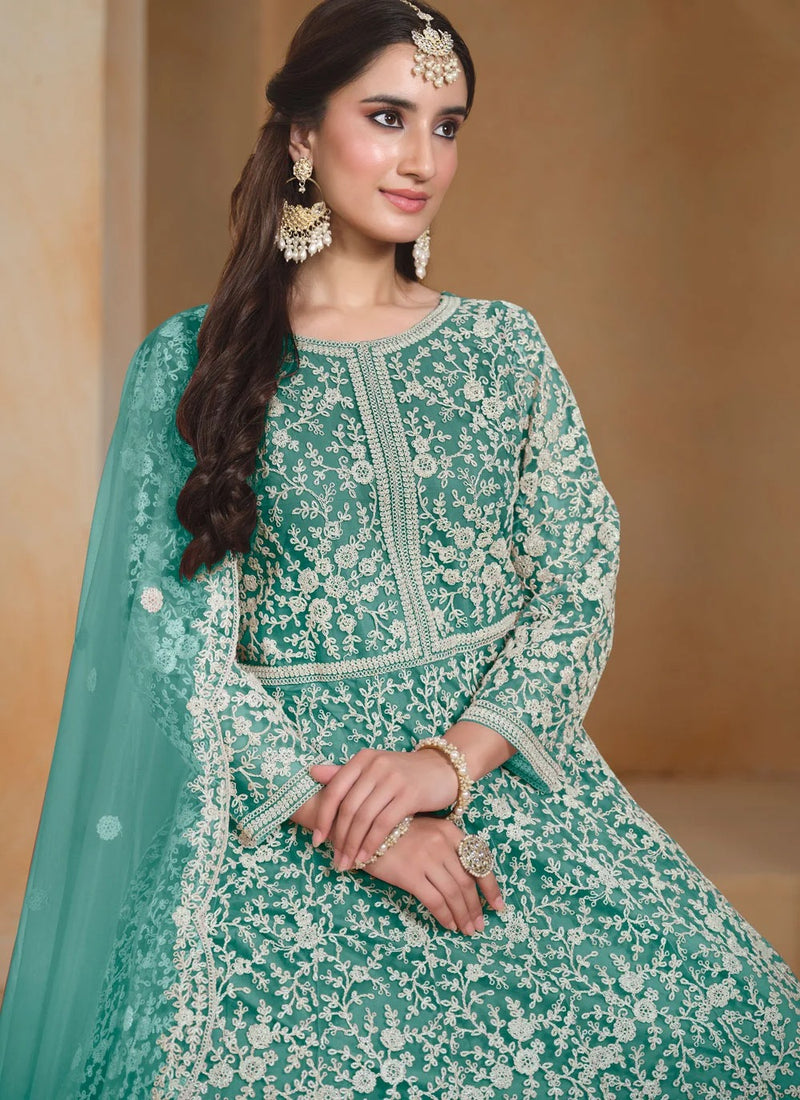 A woman wearing a mint green traditional embroidered outfit with white floral lace patterns, a matching dupatta, and ornate gold jewelry, posed against a warm beige background.