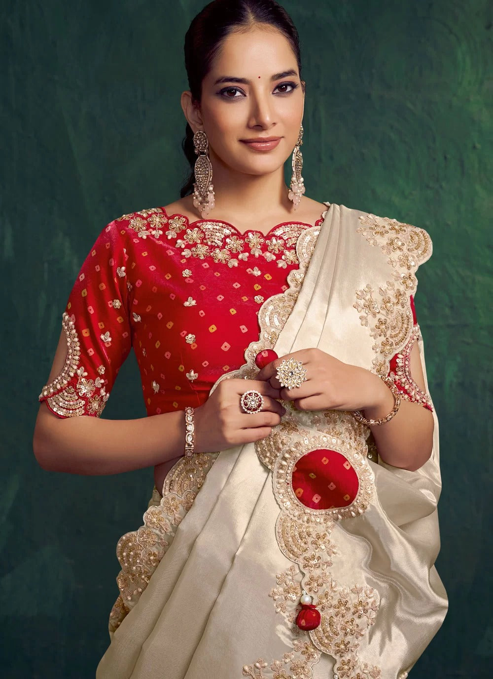 Woman wearing a red blouse with gold embroidery and a cream saree with red details against a green background