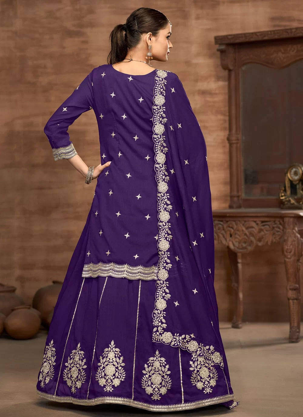 Woman wearing a purple traditional outfit with white embroidery in a room with wooden furniture.